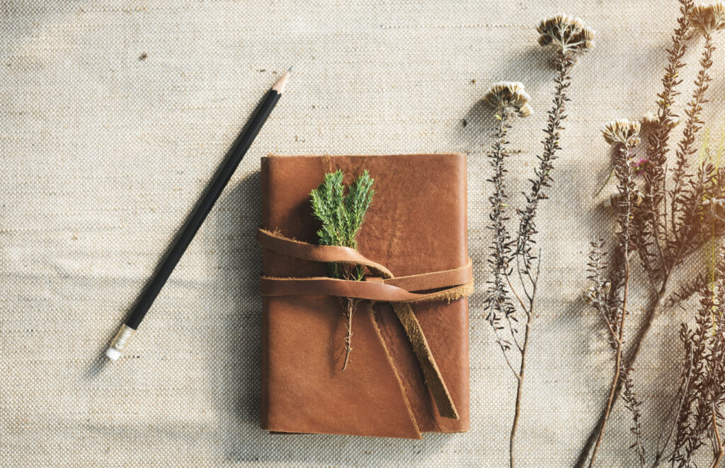 A leather - bound journal for garden notes surrounded by flowers and a pencil placed close by