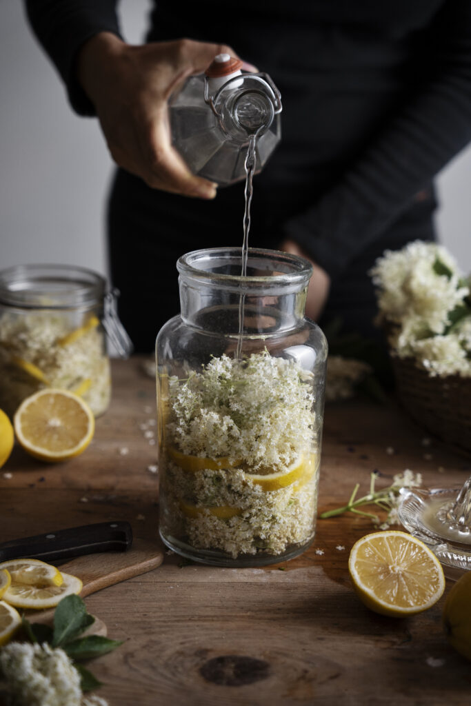 A table of jars for preserving and a person pouring liquid into a jar of elderflower and lemon making elderflower wine or champagne