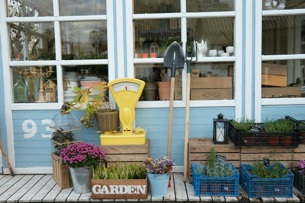 A shabby chic image which could be a garage sale, garden centre, some ones back yard or garden, with crates, garden tools, plants and a traditional weighing scale.