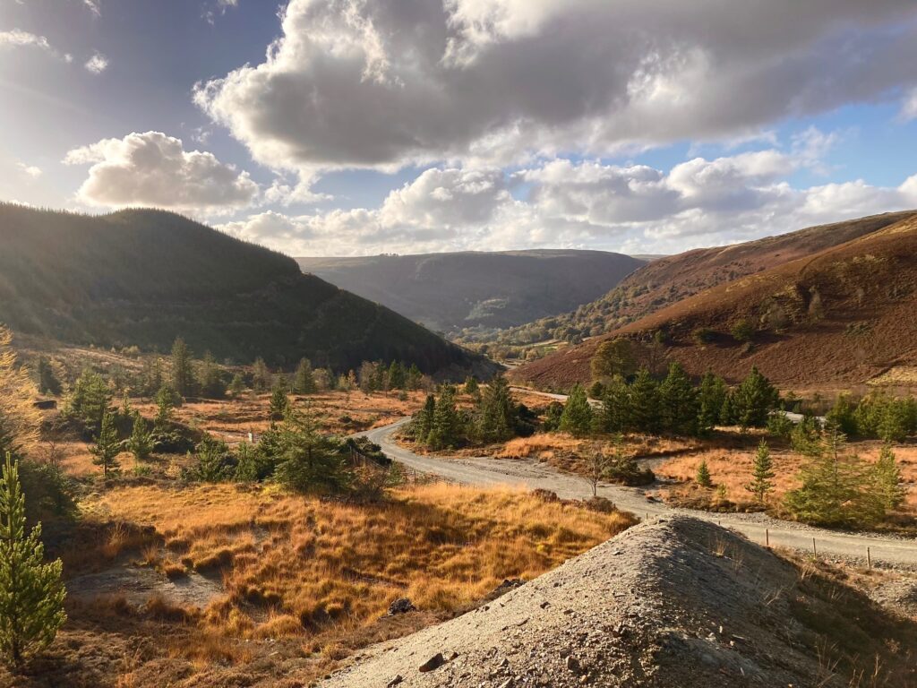 a landscape scene with mountains, brown autumn coloured grasses and a grey contrasting path with blue skies and fluffy white clouds  showing the journey to living off grid and self sufficiency